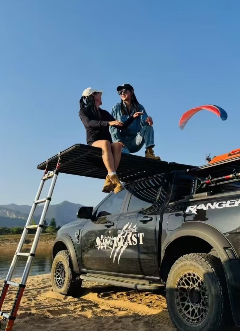 Pickup truck with overland roof rack platform and ladder on the beach, two people sitting on the rack